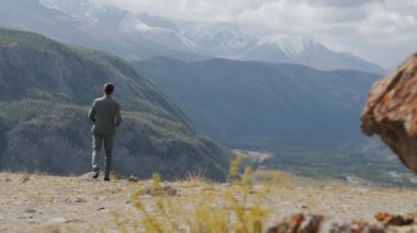 Genç bir adam dağlarda güneş gözlüğü takıyor. Bir adam dağlarda yürüyor, sırtından vurulmuş..