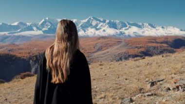 Siyah paltolu ve siyah çizmeli bir kadın dağın tepesinde yürüyor. Güneş gözlüklü genç bir kız, dağların arkasında..