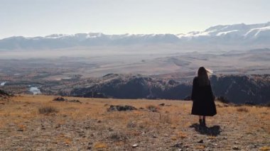 Siyah paltolu ve siyah çizmeli bir kadın dağın tepesinde yürüyor. Kız dağların arka planında.