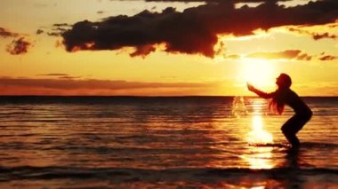 Gün batımında sahilde yürüyen bir kadın. Gün batımında suda yürüyen bir kızın silueti. Gün batımında denizde bir kız. Gün batımında ellerden su damlaları.