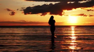 Gün batımında sahilde yürüyen bir kadın. Gün batımında suda yürüyen bir kızın silueti. Gün batımında denizde bir kız. Gün batımında ayaktan su damlaları.
