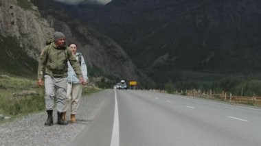 Bir kadın ve büyük turist sırt çantalı bir adam yol boyunca yürüyorlar, kaldırılmış bir el ile arabayı durdurmaya çalışıyorlar. Otostopçular, turistler yol kenarındaki dağlarda.