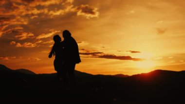 Dağlarda yaşayan evli bir çiftin siluetleri. Bir erkek ve bir kadın, dağlarda gün batımının arka planında sarılıp öpüşüyorlar. Düğün çifti. Mutlu bir aile.. 