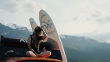 Kız, bir dağ gölünün arka planına ve yüksek dağlara karşı bir off-road aracında oturuyor, saçını düzleştiriyor. Bir kız SUP kaykayına bindikten sonra dinleniyor. Bir kız arabanın kaputunda oturuyor.. 