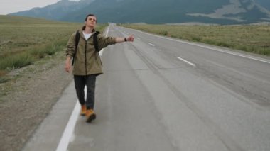 Sırt çantalı yakışıklı adam dağlarda yoldan geçen arabaları durdurmaya çalışıyor. Otostopçu adam tepe boyunca yolda. Gezgin, baş parmak kaldırarak el ele tutuşuyor.. 