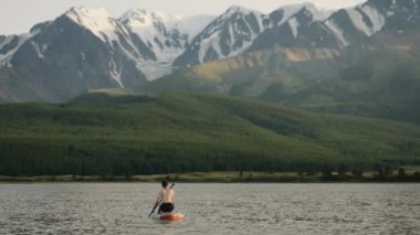 Genç adam bir dağ gölünde SUP tahtasının üzerinde yüzüyor. Rahatla, Faaliyet, SUP Biniş, Stand Up Spor Eğlencesi Kürekçiliği.