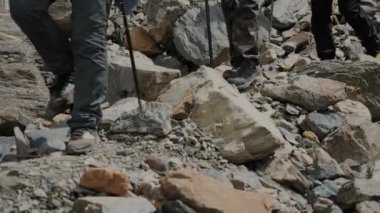 Yürüyüşçülerin bacaklarının yürüyüş direğiyle yakın çekimi. Bir grup turist yürüyüş direkleriyle kayaların üzerinden dağlara doğru yürüyorlar. Dağ turizmi.