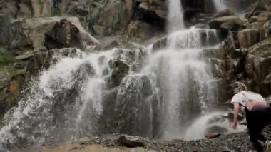 Bir turist kız bir şelaleye tırmandı. Bir turist, dağ şelalesindeki başarısına sevinir. Şelalenin yakınındaki dağlarda bir kız turist.