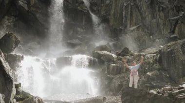 Bir turist kız bir şelaleye tırmandı. Bir turist atlıyor ve onun dağ şelalesindeki başarısına seviniyor. Şelalenin yakınındaki dağlarda bir kız turist.