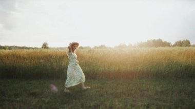 Gün batımında tarlada yürüyen elbise ve şapkalı güzel bir kızın portresi. Kameraya bakıyor ve gülümsüyor..