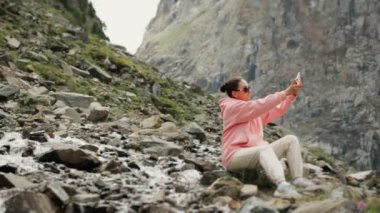 Güneş gözlüklü ve pembe kazaklı bir turist dağlarda kayalara oturup telefonuyla güzel dağ manzaralarının fotoğraflarını çekiyor..