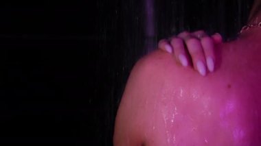 Genç bir kız sırtüstü duruyor ve neon ışıkta karanlık bir banyoda duş alıyor. Elini omzunun ve boynunun üstünden geçiriyordu. Vücut bakımı ve güzellik konsepti.