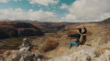 Sırtında sırt çantası olan bir erkek turist dağın tepesine oturur ve bir taş atar. Uçurumun kenarında sırt çantalı bir turist. Seyahat ve dağ turizmi konsepti. Vahşi doğada yürüyüş..