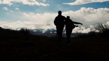 Birbirine aşık iki turistin siluetleri, bir adam ve bir kadın, birbirine sarılıp dağın zirvesine doğru yürüyorlar. Bir erkek bir kıza sarılır. Seyahat ve dağ turizmi konsepti. Vahşi doğada yürüyüş..