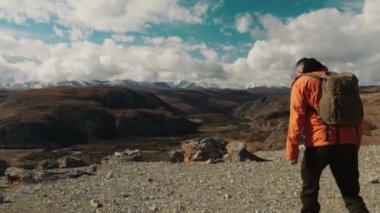 Omuzlarında sırt çantası olan turuncu ceketli bir erkek turist dağın zirvesine çıkar ve ellerini kaldırarak başarısını kutlar. Seyahat ve turizm kavramı.