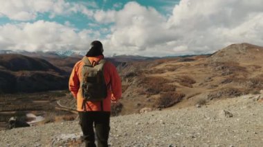 Omuzlarında sırt çantası olan turuncu ceketli bir erkek turist dağlarda yürüyor. Bir turist sonbaharda dağlara seyahat eder. Seyahat ve turizm kavramı.
