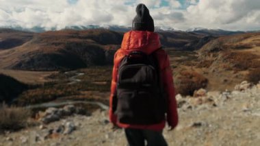Omuzlarında sırt çantası olan kırmızı ceketli bir turist kız dağlarda yürüyor. Sonbaharda, bir turist dağlarda seyahat eder. Seyahat ve turizm kavramı.