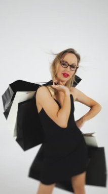 Siyah elbiseli ve alışveriş torbalı bir kız stüdyoda beyaz arka planda poz veriyor. Bir kız alışveriş torbalarıyla kameraya poz veriyor. Satış, mutlu alıcı. Mevsimlik indirimler. Dikey video.