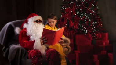 Küçük bir kız Noel Baba 'nın kucağında Noel ağacının yanındaki salonda oturuyor ve Noel kitabı okuyor. Çocuk ve Noel Baba. Noel ve yeni yılı kutlamak için konsept. Mutlu Noeller..