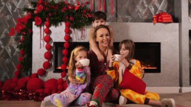 Çocuklu mutlu anne, Noel için dekore edilmiş oturma odasındaki şöminenin yanında oturuyor. Çocuklar kakao içer ve gülümser. Noel ve Yeni Yıl kutlamaları. Mutlu Noeller..
