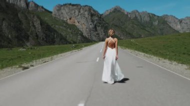 Beyaz elbiseli bir kız dağlarda yol boyunca yürür. Bir kız dağ yolunda gelinlikle yürüyor..