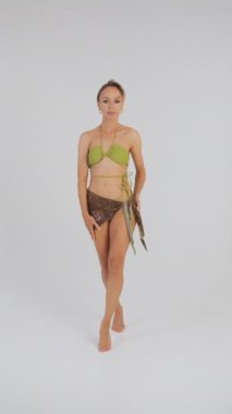 Yeşil mayo giyen ve kameraya pareo pozu veren güzel bir kız. Stüdyoda beyaz arka planda plaj kıyafeti giyen bir kız. Plaj modası. Mayolu kız. Dikey video.