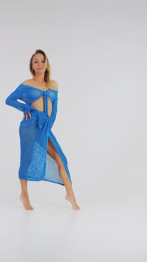 Mavi mayo giymiş genç ve güzel bir kız kameranın önünde poz veriyor ve vücudunu okşuyor. Stüdyoda beyaz arka planda plaj kıyafeti giyen bir kız. Plaj modası. Mayolu kız. Dikey video.