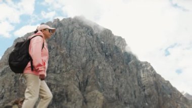 Sırtında sırt çantası ve güneş gözlükleriyle pembe ceketli bir turist kız dağların güzel manzarasına hayran. Dağlardaki kız turist.