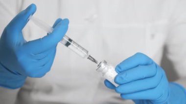 Doktorun mavi eldivenli hali, şişedeki bir maddenin iki bileşenini karıştırıyor. Araştırmacı maddeyi şırıngayla şişeye enjekte ediyor..