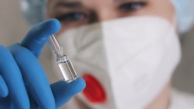 Solunum cihazlı ve koruyucu eldivenli bir kadın doktor elinde aşı olan bir ampul tutuyor. Doktor ampulü ilaçla kontrol ediyor..