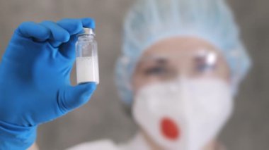 Solunum cihazlı ve koruyucu eldivenli bir doktor elinde aşı olan bir ampul tutuyor ve çerçeveye bakıyor. Doktor elinde ilaçla bir ampul tutuyor..