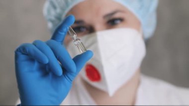 Solunum cihazlı ve koruyucu eldivenli bir kadın doktor elinde aşı olan bir ampul tutuyor. Doktor ampulü ilaçla kontrol ediyor..