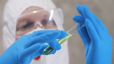Koruyucu kıyafetli, gözlüklü ve mavi eldivenli bir bilim adamı elinde numune olan bir test tüpü tutuyor. Tıbbi laboratuvar. Bilimsel Laboratuvar.