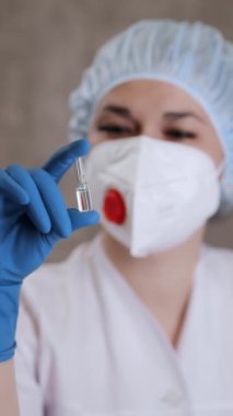 Solunum cihazlı ve koruyucu eldivenli bir kadın doktor elinde aşı olan bir ampul tutuyor. Doktor ampule ilaçla bakıyor. Dikey video.