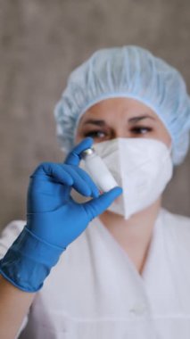Solunum cihazlı ve koruyucu eldivenli bir kadın doktor bir ampulü aşıyla sallıyor. Doktor ampulü ilaçla sallıyor. Dikey video.