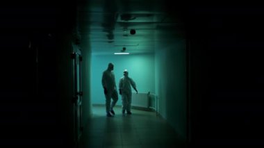 Koruyucu takım elbiseli iki doktor karanlık bir koridor boyunca bir hastanede yürüyor. Kimyasal koruyucu elbiseli iki kişi koridorda yürüyor..