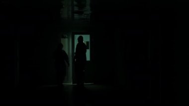 Koruyucu takım elbiseli iki doktor hastane karanlık bir koridor boyunca yürür, kapıyı açar ve içeri girerler. Kimyasal koruyucu elbiseli iki kişi koridorda yürüyor..