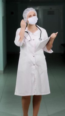 Elinde bir pano olan, elinde koruyucu bir maske olan ve gülümseyen ve kameraya bakan bir kadın doktor portresi. Dikey video.