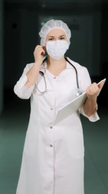 Elinde bir pano olan, elinde koruyucu bir maske olan ve gülümseyen ve kameraya bakan bir kadın doktor portresi. Dikey video.