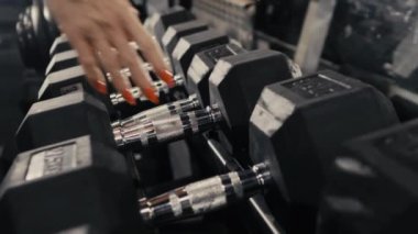 Bir kadının ellerinin raftan dambıl alırken yakın plan çekimi. Spor malzemeleri. Spor salonu. Dambıllı kol eğitimi.