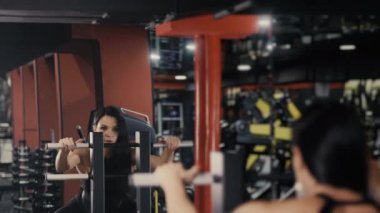 Genç bir sporcu kız, spor salonunda idman yapıyor ve bir makineye ağırlık verip çömeliyor. Sırtını ve bacaklarını güçlendirmek için egzersiz yap. Spor salonunda kız eğitimi. Modern spor salonu.