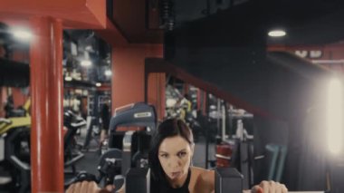 Bir kız sporcu egzersiz yapıyor, egzersiz makinesinin saplarını tutuyor ve kameraya bakıyor. Spor salonunda kız eğitimi..