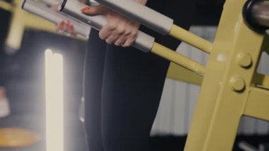 Egzersiz makinesinin kulplarını tutan bir kızın yakın çekimi. Genç bir sporcu kız, spor salonunda idman yapıyor ve bir makineye ağırlık verip çömeliyor. Spor salonunda kız eğitimi..