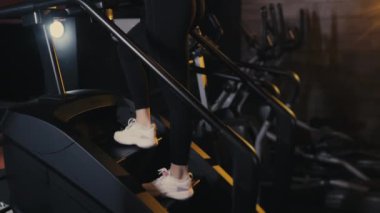 Egzersiz makinesinin basamaklarında yürüyen bir kız sporcunun bacaklarına yakın çekim. Spor üvey antrenörü. Bir kız spor salonunda antrenman yapıyor. Merdiven tırmanma egzersiz makinesi.