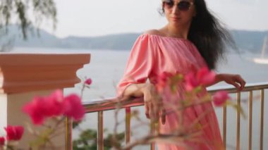 Pembe elbiseli ve güneş gözlüklü genç bir kızın portresi okyanusa bakan bir odanın balkonunda kameraya poz veriyor. Kız turist, tropik bir adada tatilde..