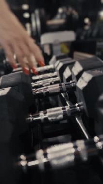 Bir kadının ellerinin raftan dambıl alırken yakın plan çekimi. Spor malzemeleri. Spor salonu. Dambıllı kol eğitimi. Dikey video.