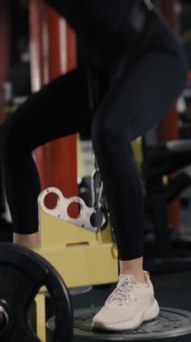 Genç bir sporcu kız, spor salonunda idman yapıyor ve bir makineye ağırlık verip çömeliyor. Sırtını ve bacaklarını güçlendirmek için egzersiz yap. Spor salonunda kız eğitimi. Modern spor salonu. Dikey video.