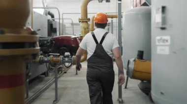 Kasklı ve üniformalı bir işçi elinde İngiliz anahtarıyla kazan dairesinde gaz boruları ve kazanlar boyunca yürüyor. İş üniformalı mühendis..
