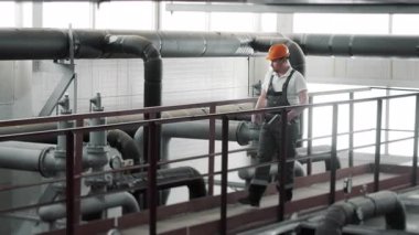 Güvenlik kaskı ve üniforma mühendisi elinde tabletle fabrikadaki boru bağlantısını kontrol ediyor ve notlar alıyor. İş üniformalı bir mühendis. Bakım ve izleme.