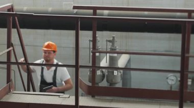 Elinde tabletle kasketli ve üniformalı bir mühendis, ekipmanları kontrol etmek için fabrikada metal endüstriyel bir merdivene tırmanıyor. Üniformalı fabrika çalışanı..